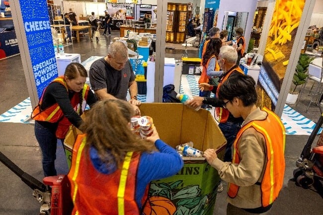 People in safety vests putting various items into a large cardboard box at an event.