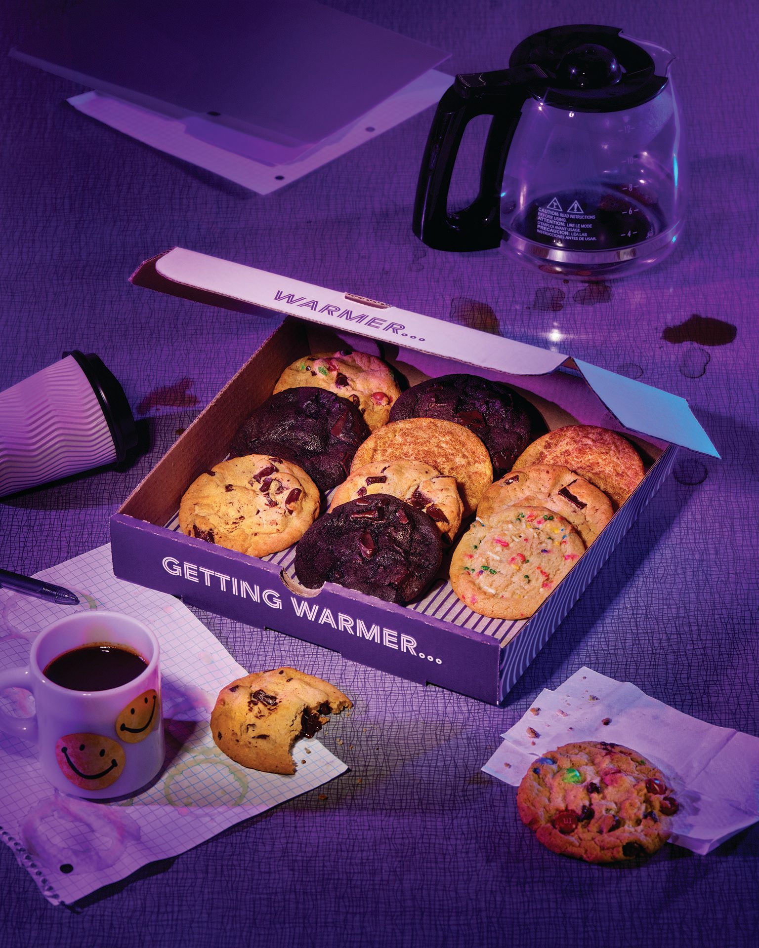 An open box of assorted cookies, coffee, and spills on a purple-lit desk.