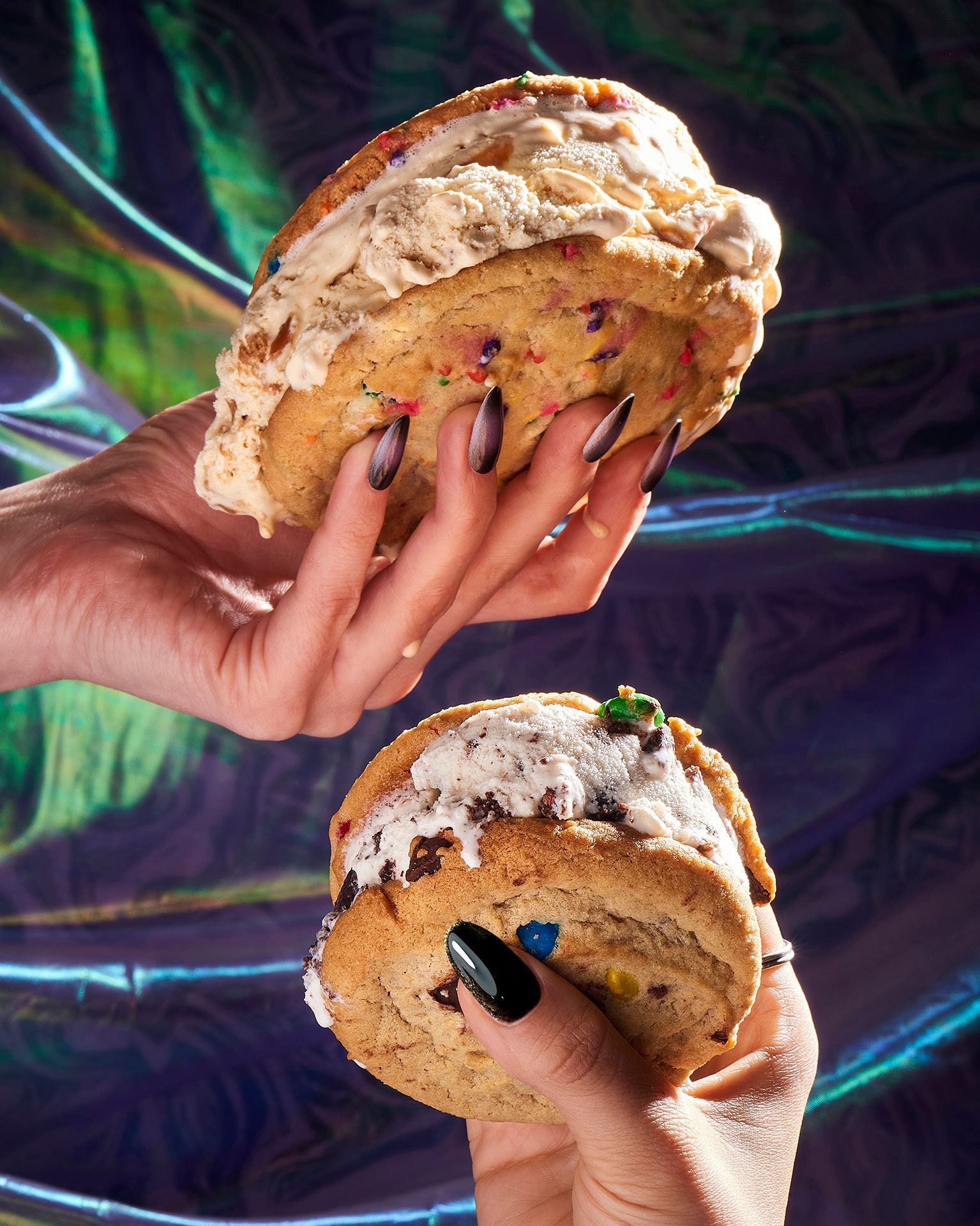Two hands with pointed dark nails hold melting ice cream sandwiches, against an iridescent background.