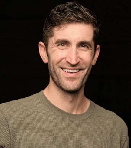 Headshot of a smiling man with short hair and a beard wearing a green shirt against a black background.