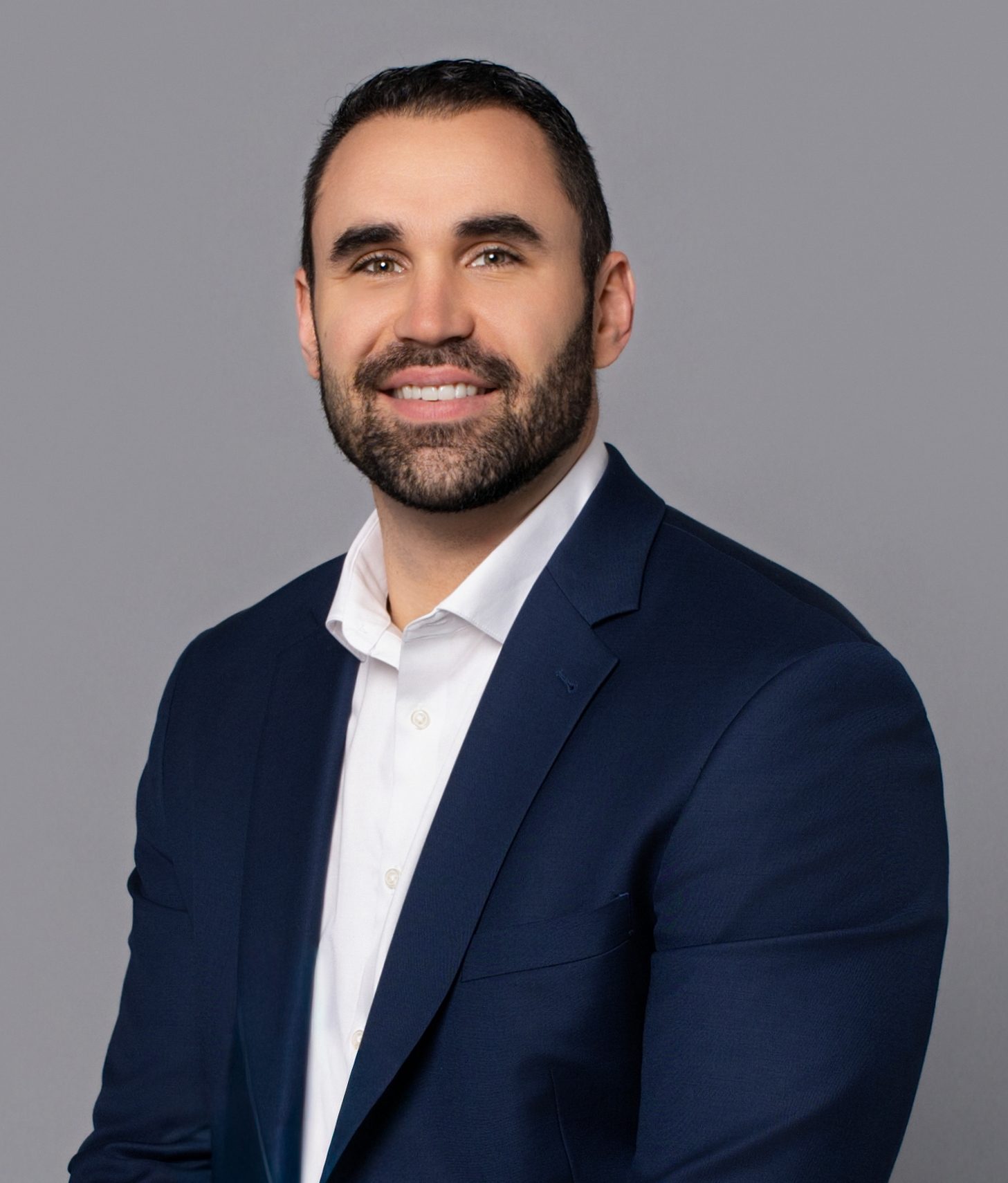 A smiling man with a beard in a dark blue suit jacket and white shirt against a gray background.