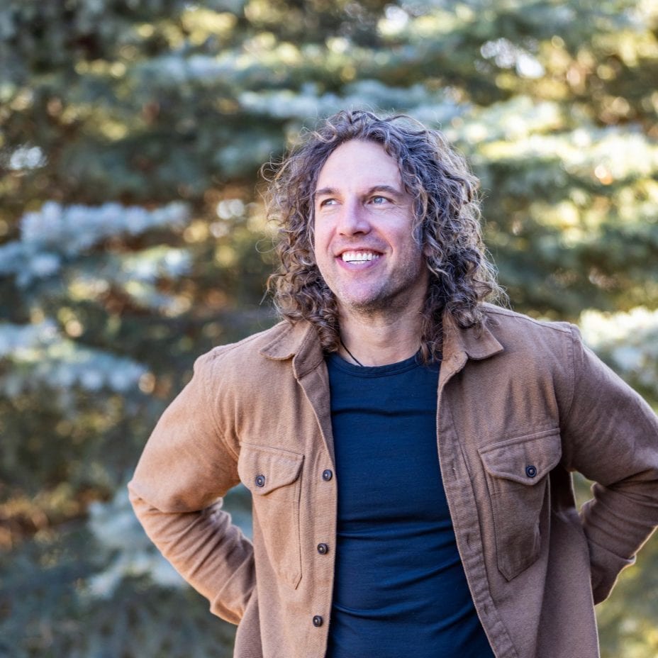 Smiling man with long curly hair and brown jacket looking up outdoors.