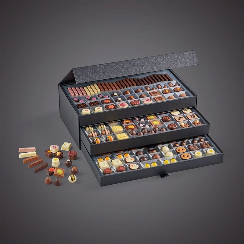 A large, three-tiered open box filled with assorted chocolates, with more chocolates scattered nearby.