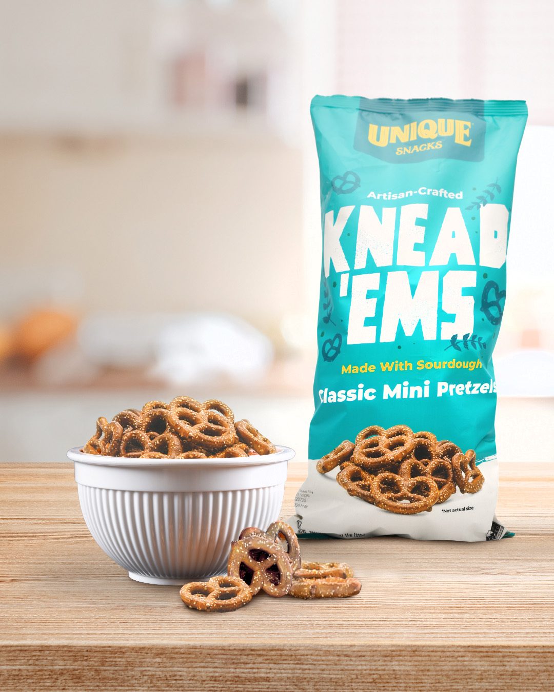 A bag of Unique Snacks Knead 'Ems pretzels next to a bowl of pretzels on a wooden table.
