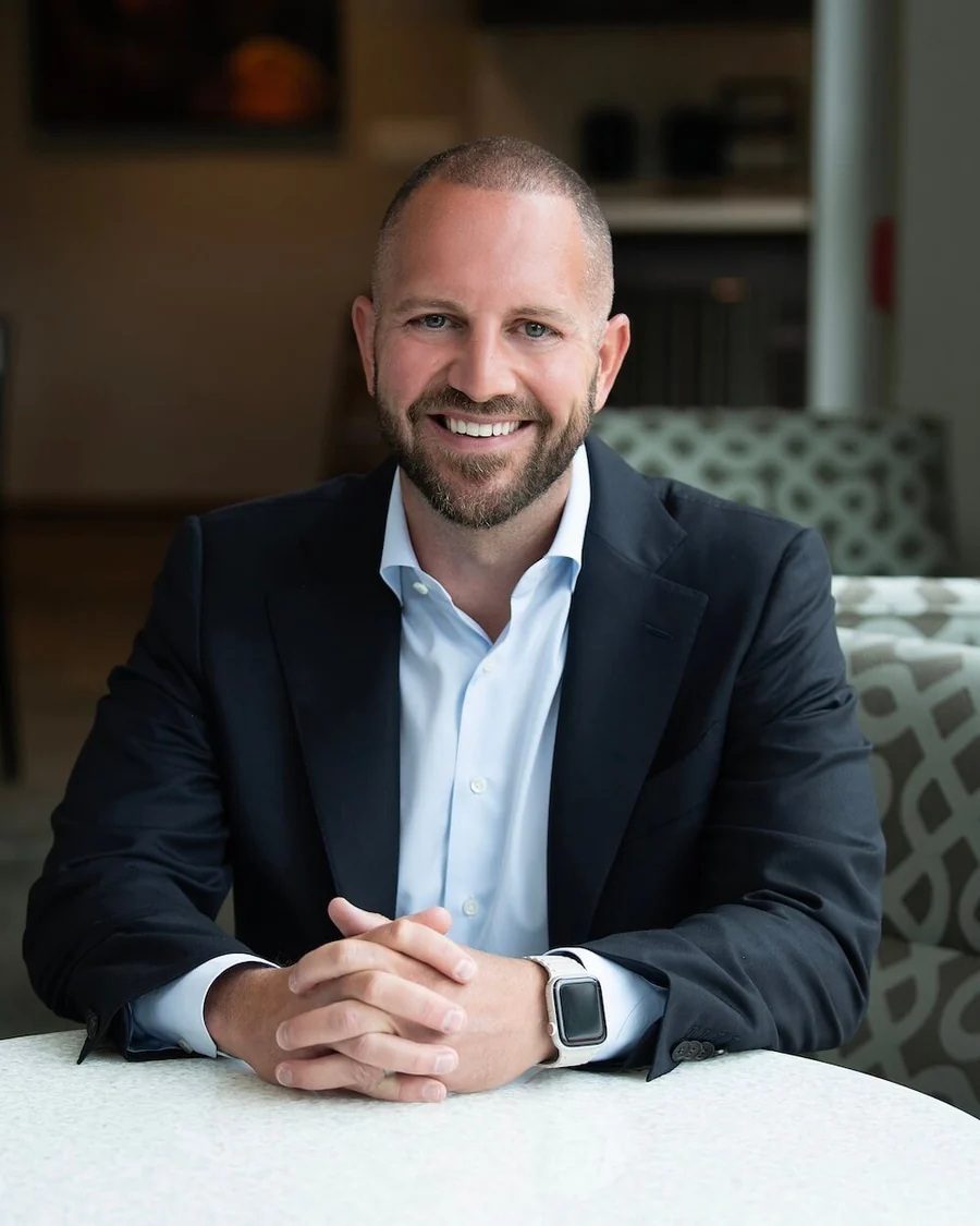 A smiling man with a beard and short hair, wearing a suit and light blue shirt, sitting at a table.