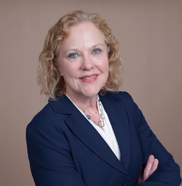 A smiling woman with curly blonde hair, wearing a dark blue suit jacket and white blouse, arms crossed.