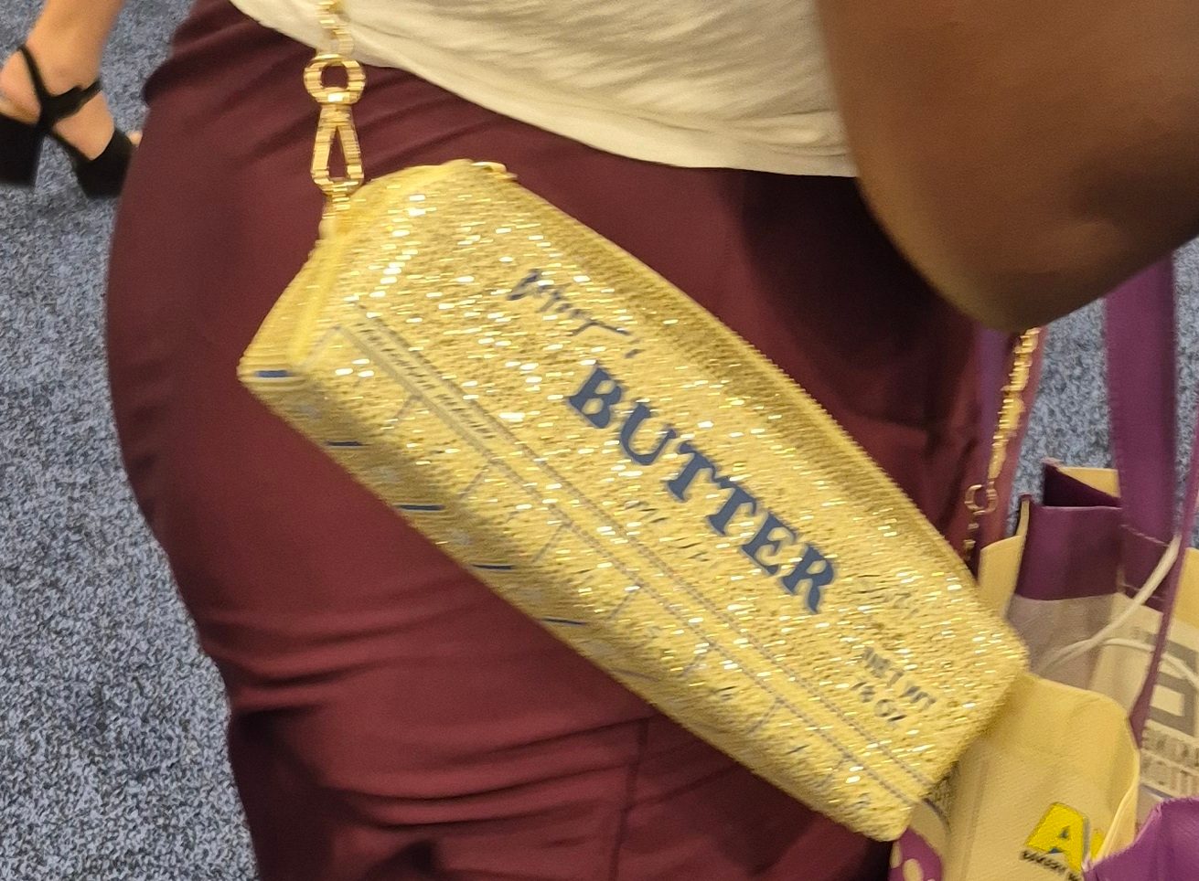 A person wears a glittery golden clutch purse shaped like a stick of butter, with "BUTTER" written on it.