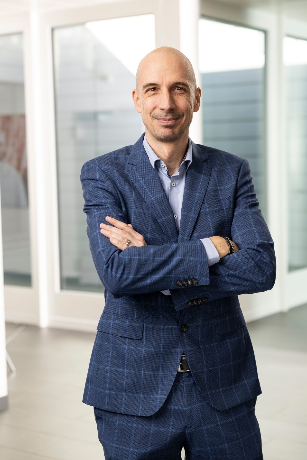 Smiling bald man in a blue plaid suit stands with crossed arms in an office setting.