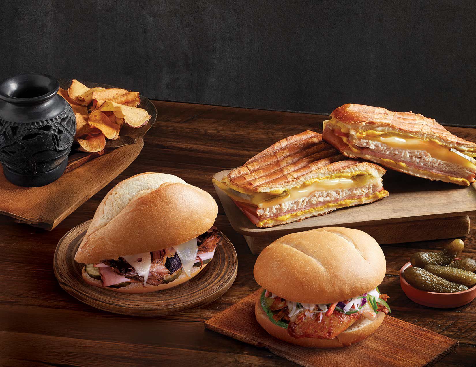 Four sandwiches (Cuban paninis, pulled pork, chicken), chips, and pickles on wood.