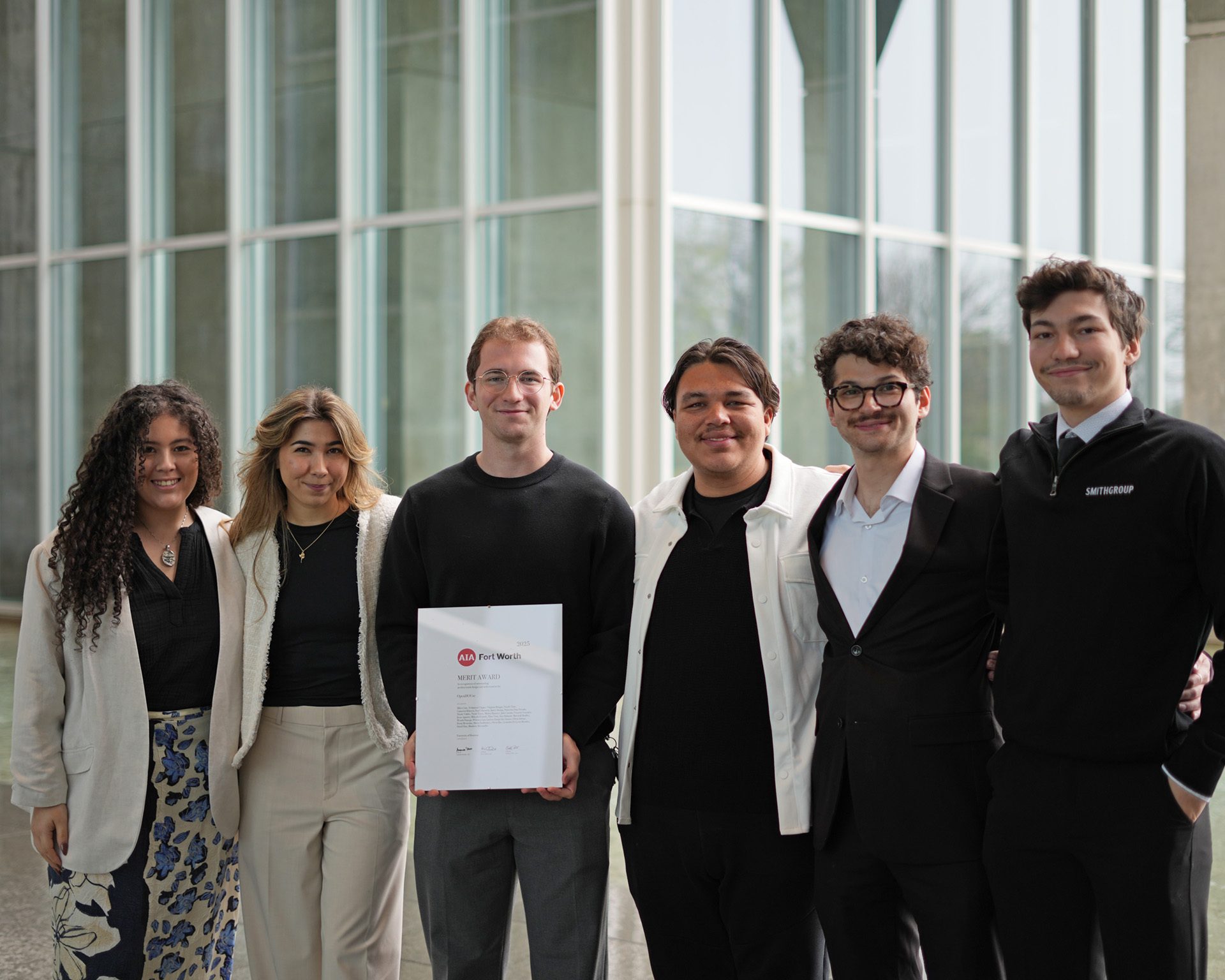 Six young adults, three men and three women, pose with an award in front of a modern building.