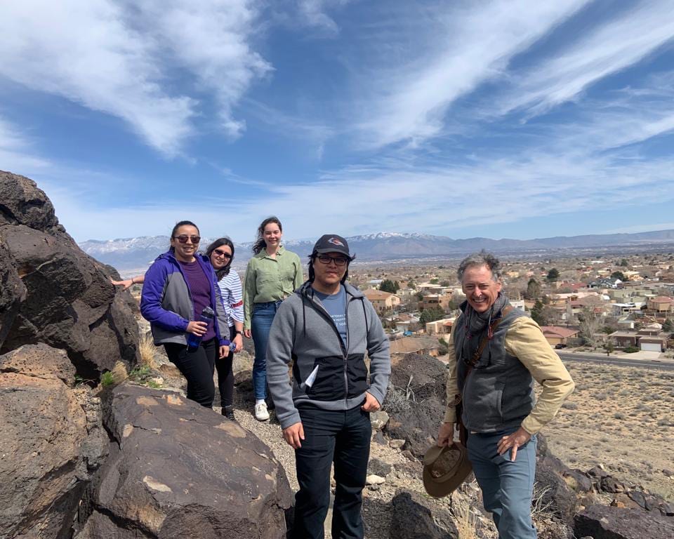 Five people on a rocky hill overlooking a city with snowy mountains and a cloudy sky.