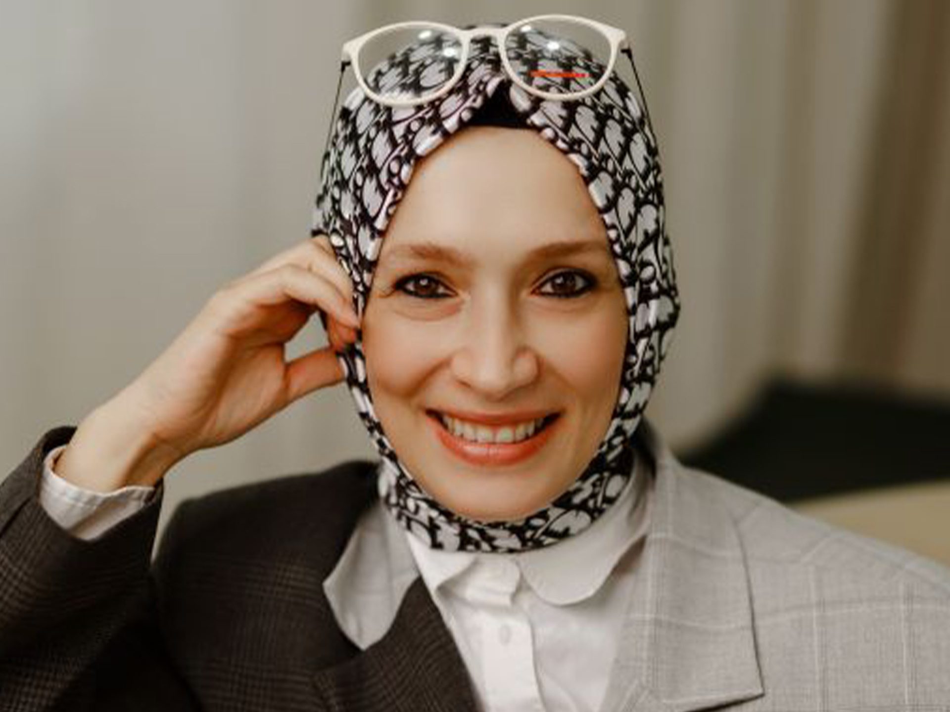 Smiling woman in patterned hijab, white glasses on top, two-toned blazer.