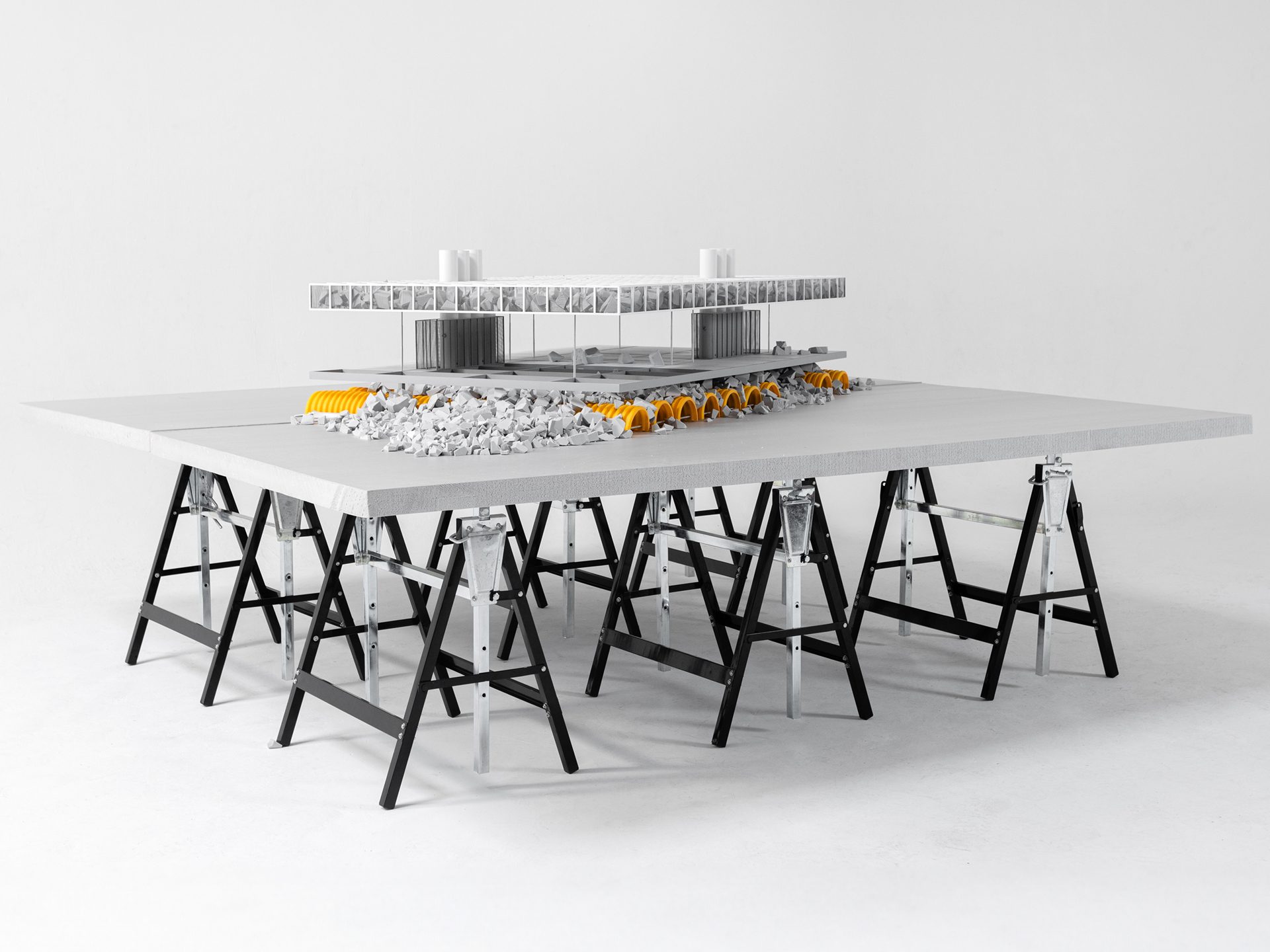 Architectural model of a building with rubble, orange tunnels, on a grey table with black sawhorses.
