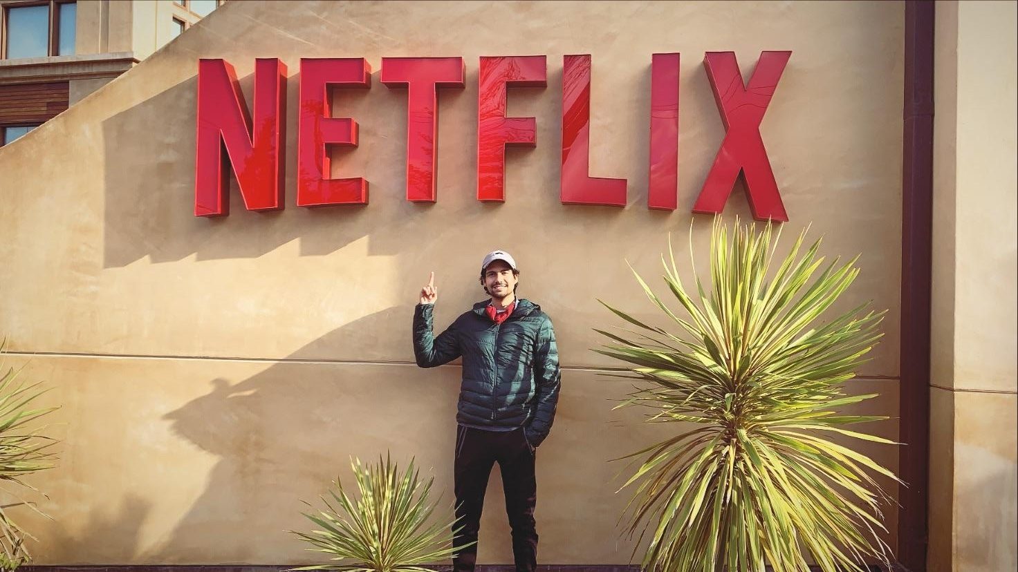 Man pointing at a large red "NETFLIX" sign on a wall, with plants.