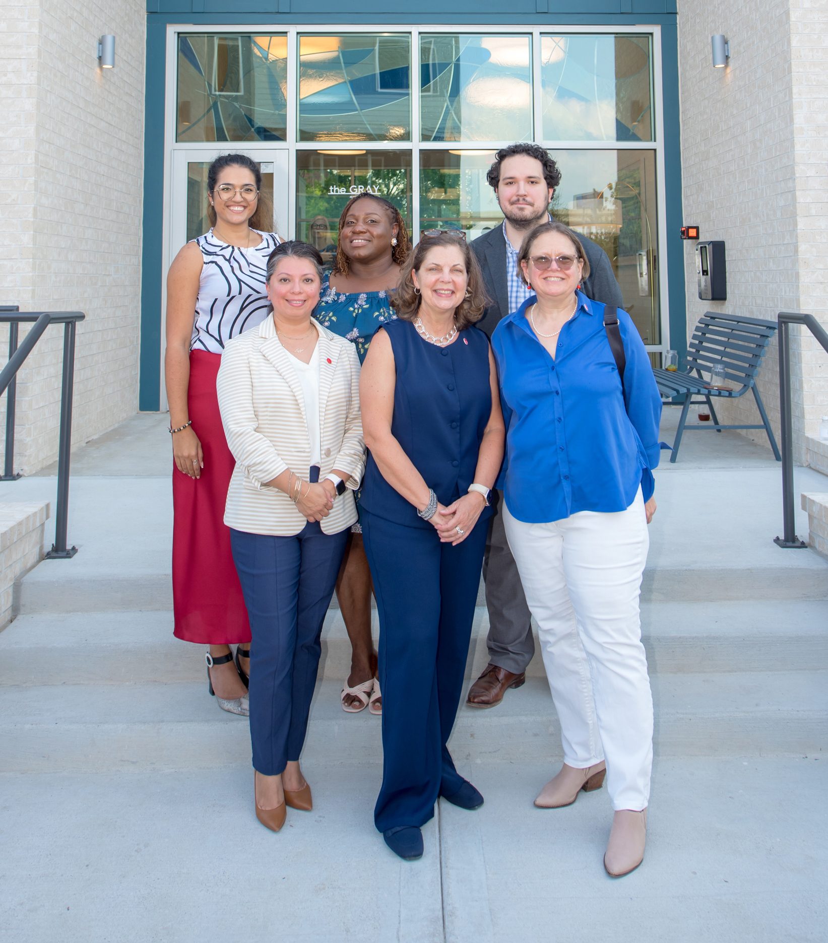 Group of six diverse individuals smiling on steps outside a modern building.