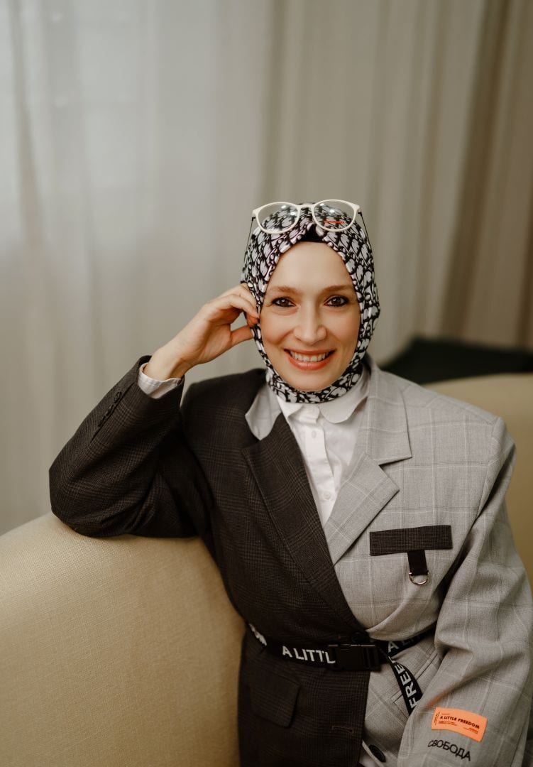 Smiling woman in patterned hijab with glasses, wearing an asymmetrical blazer, on a sofa.