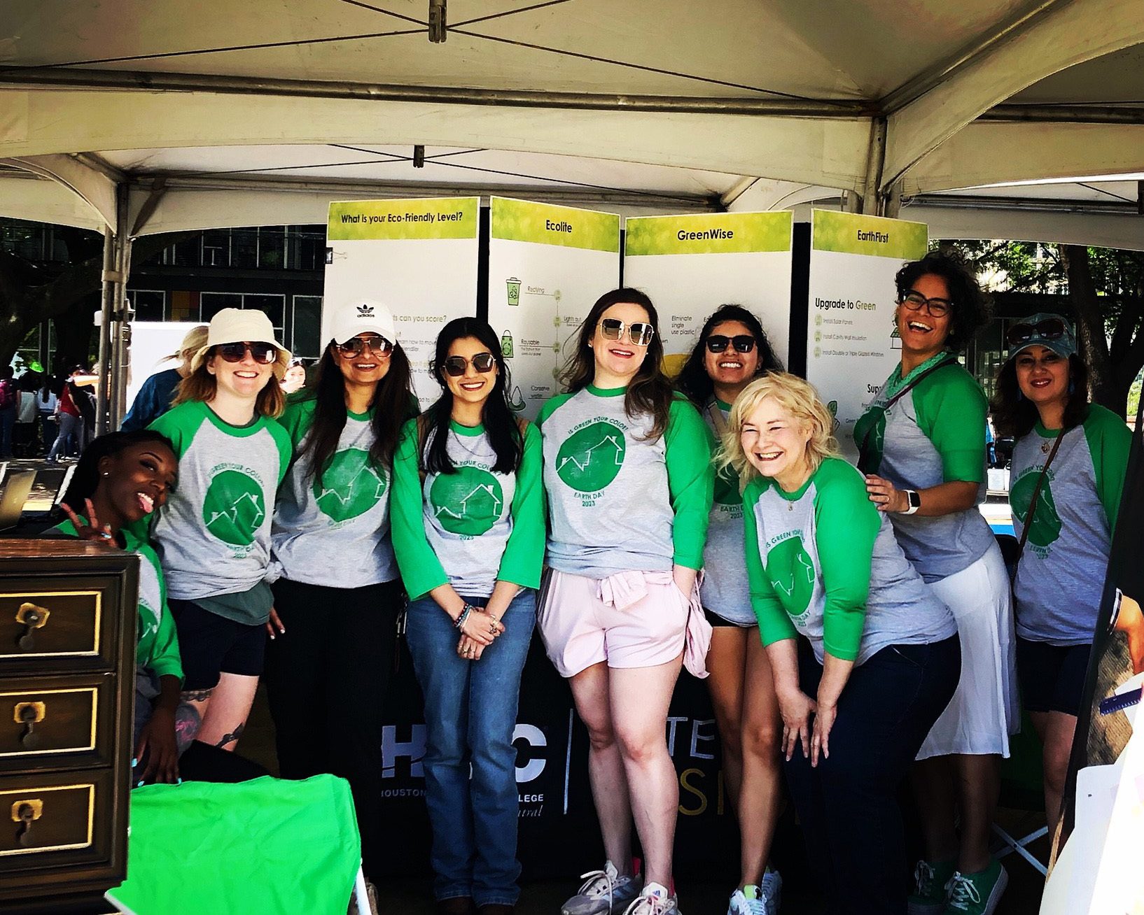 A diverse group of women in green and grey Earth Day shirts posing happily at an outdoor event.