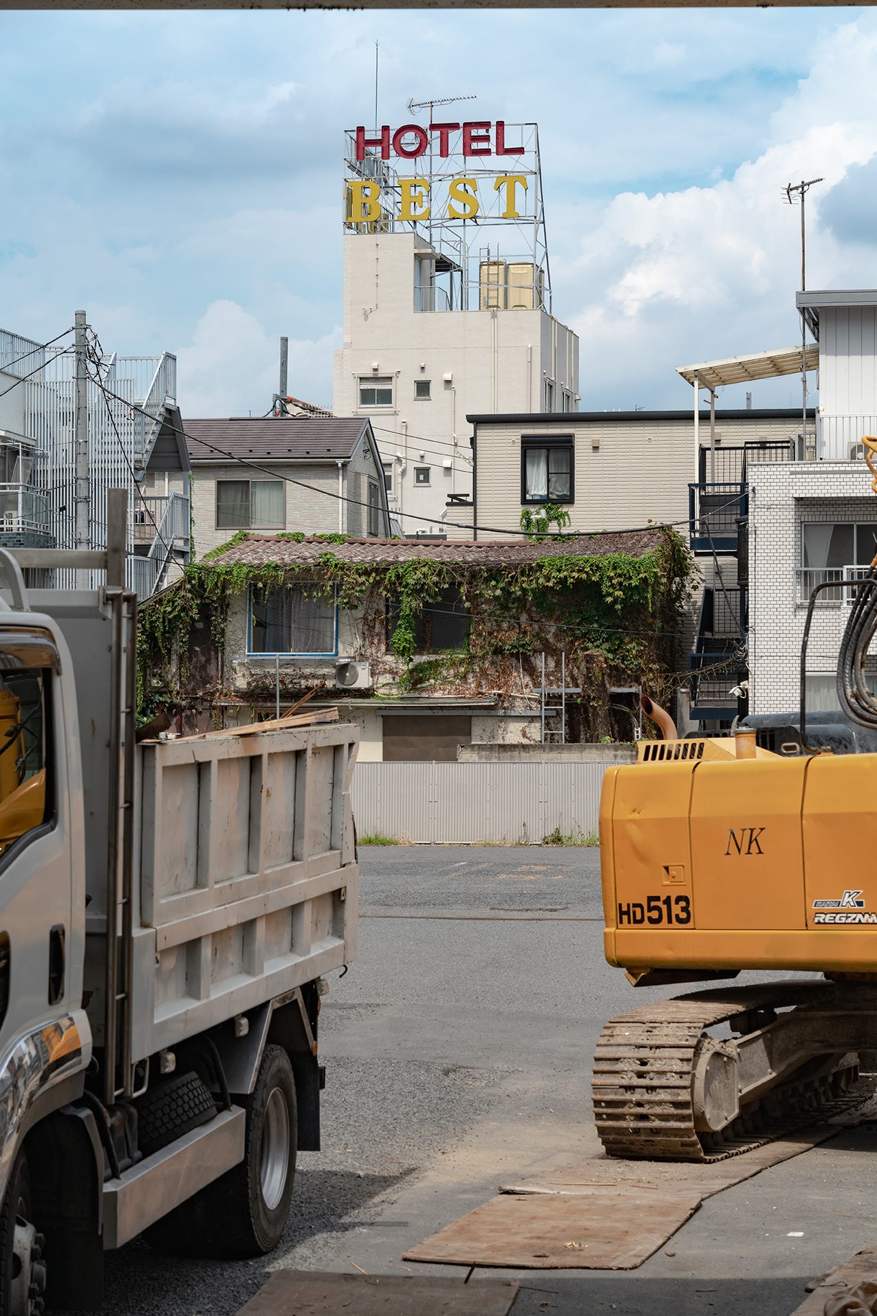 "Hotel Best" sign above vine-covered buildings, with a dump truck and excavator in the foreground.