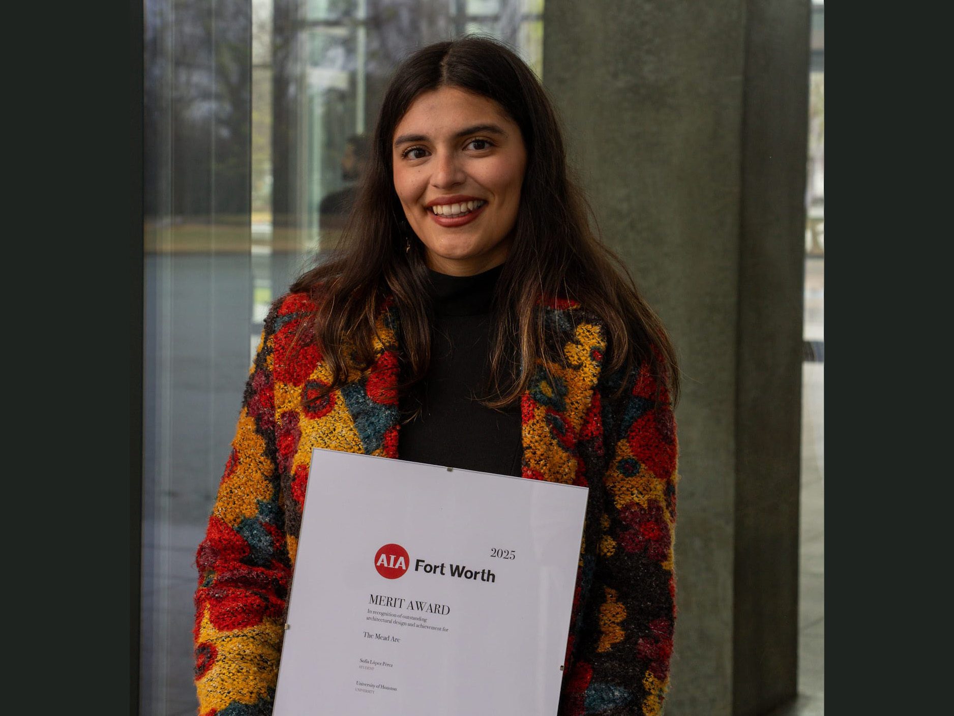 Smiling woman in a colorful jacket holding an AIA Fort Worth Merit Award certificate.