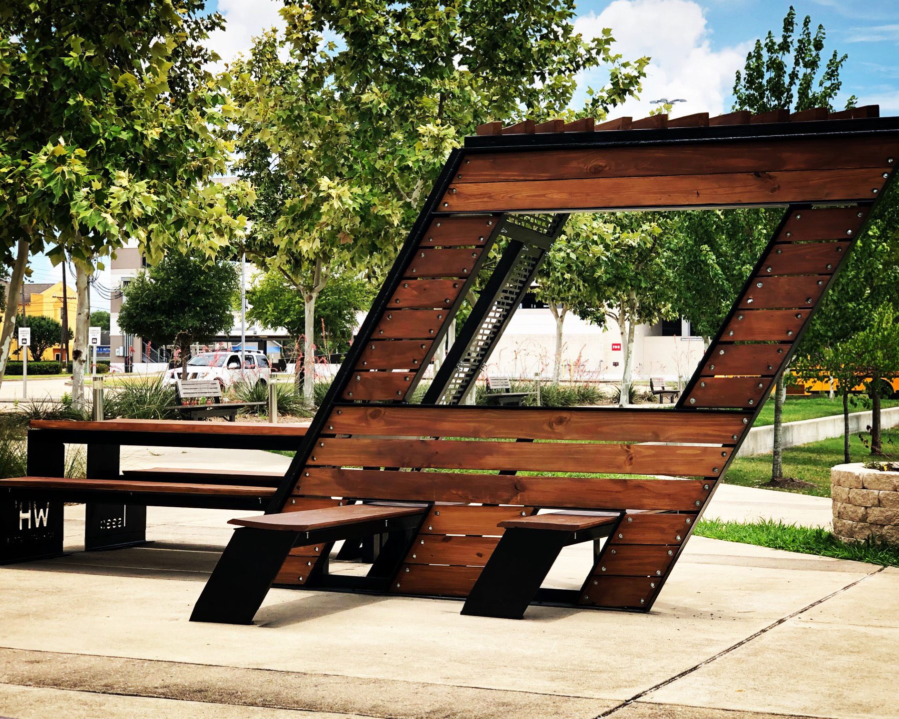 An innovative angular wooden and metal public seating structure with integrated table amidst green trees.