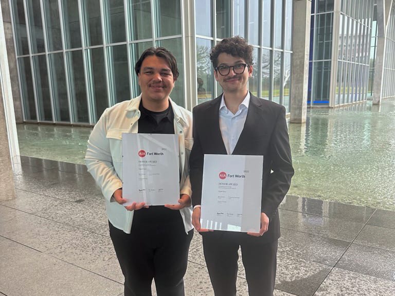 Two men proudly stand holding their AIA Fort Worth Honor Awards indoors, with a modern building and water feature.