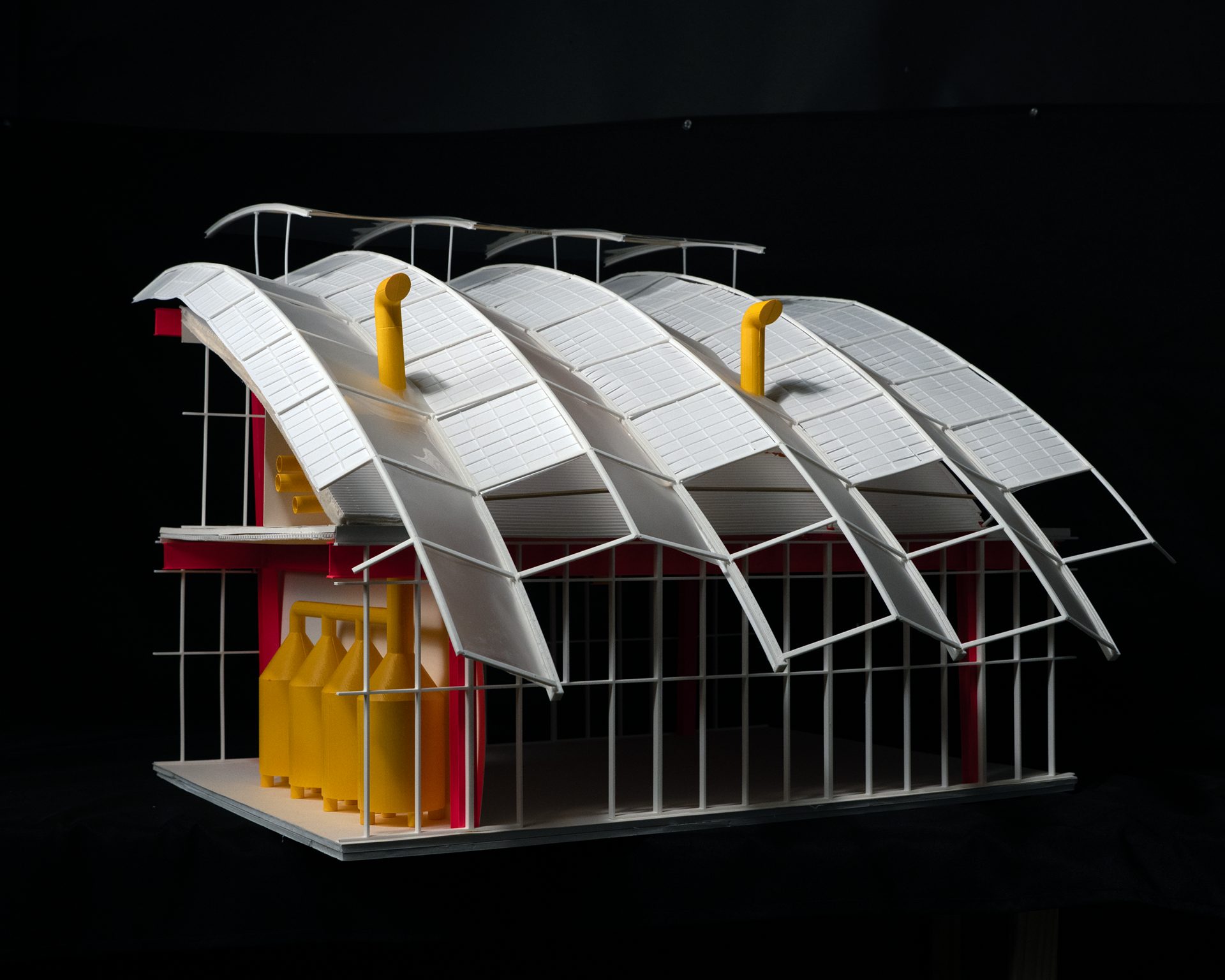 A detailed model of a factory building with a curved white roof, red frame, and yellow industrial tanks.