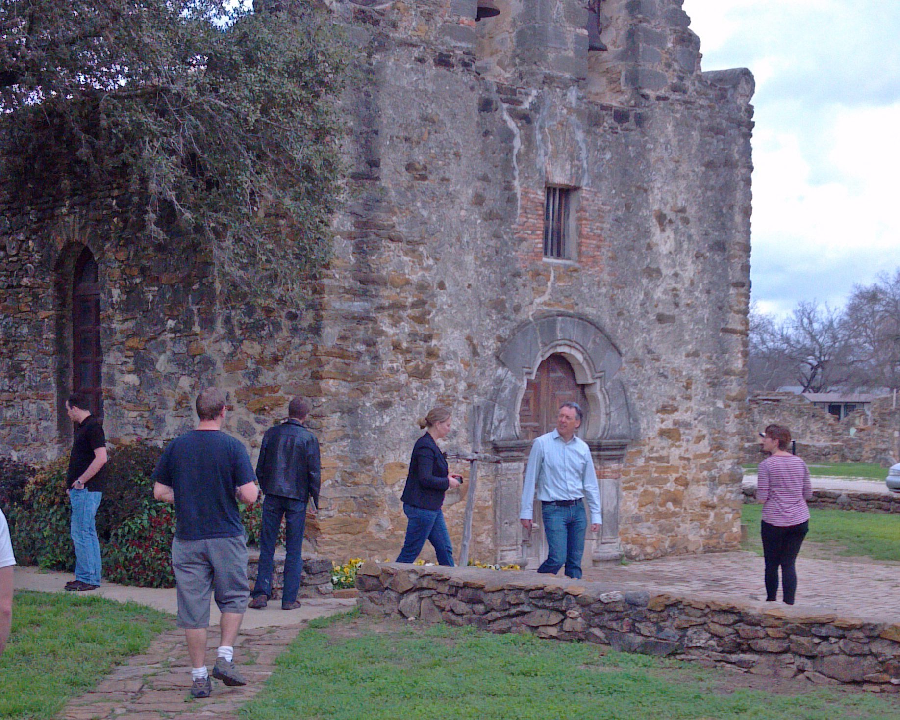People exploring an ancient stone mission building with trees.