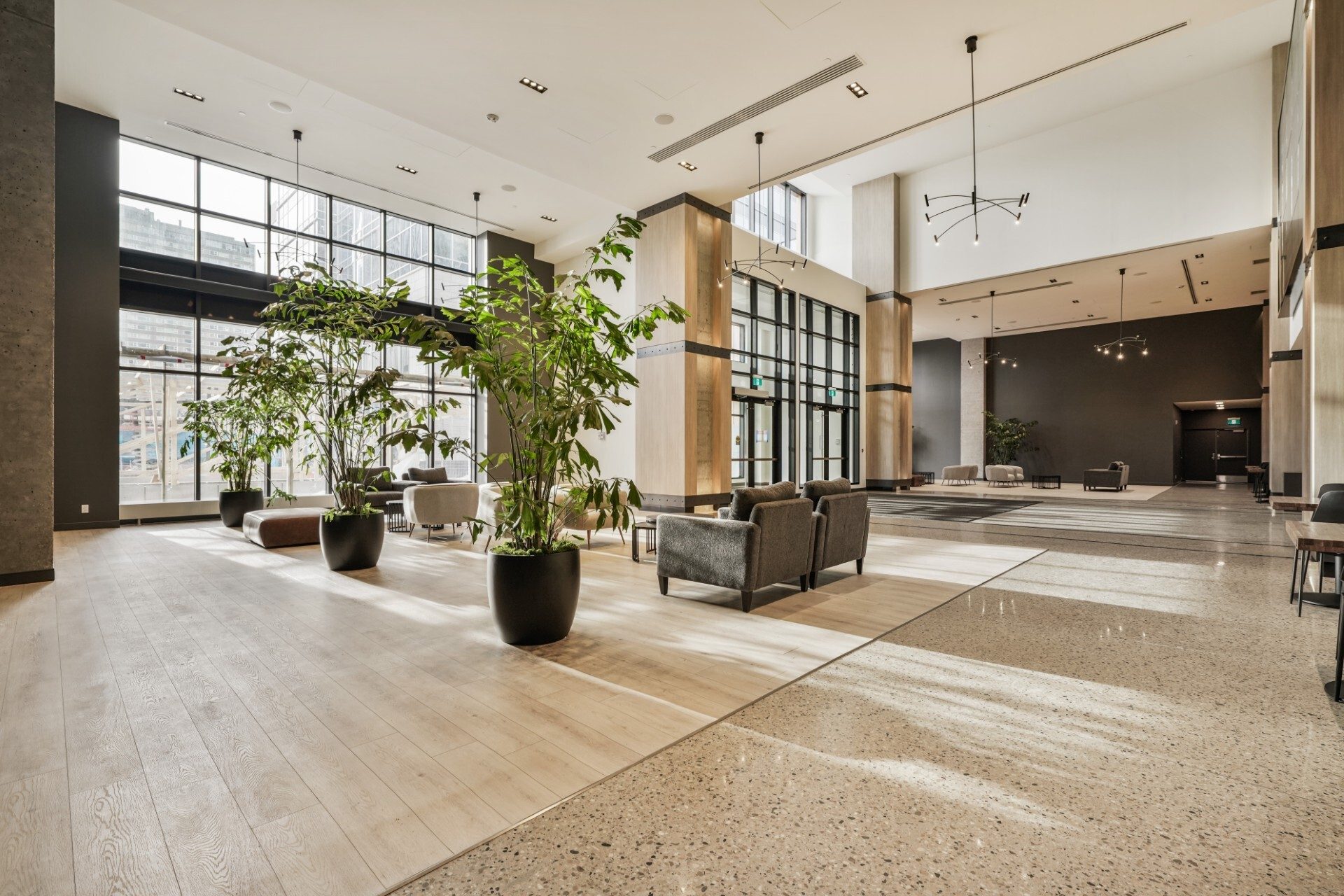Large, bright modern lobby with tall windows, potted plants, various seating, and mixed wood/terrazzo flooring.