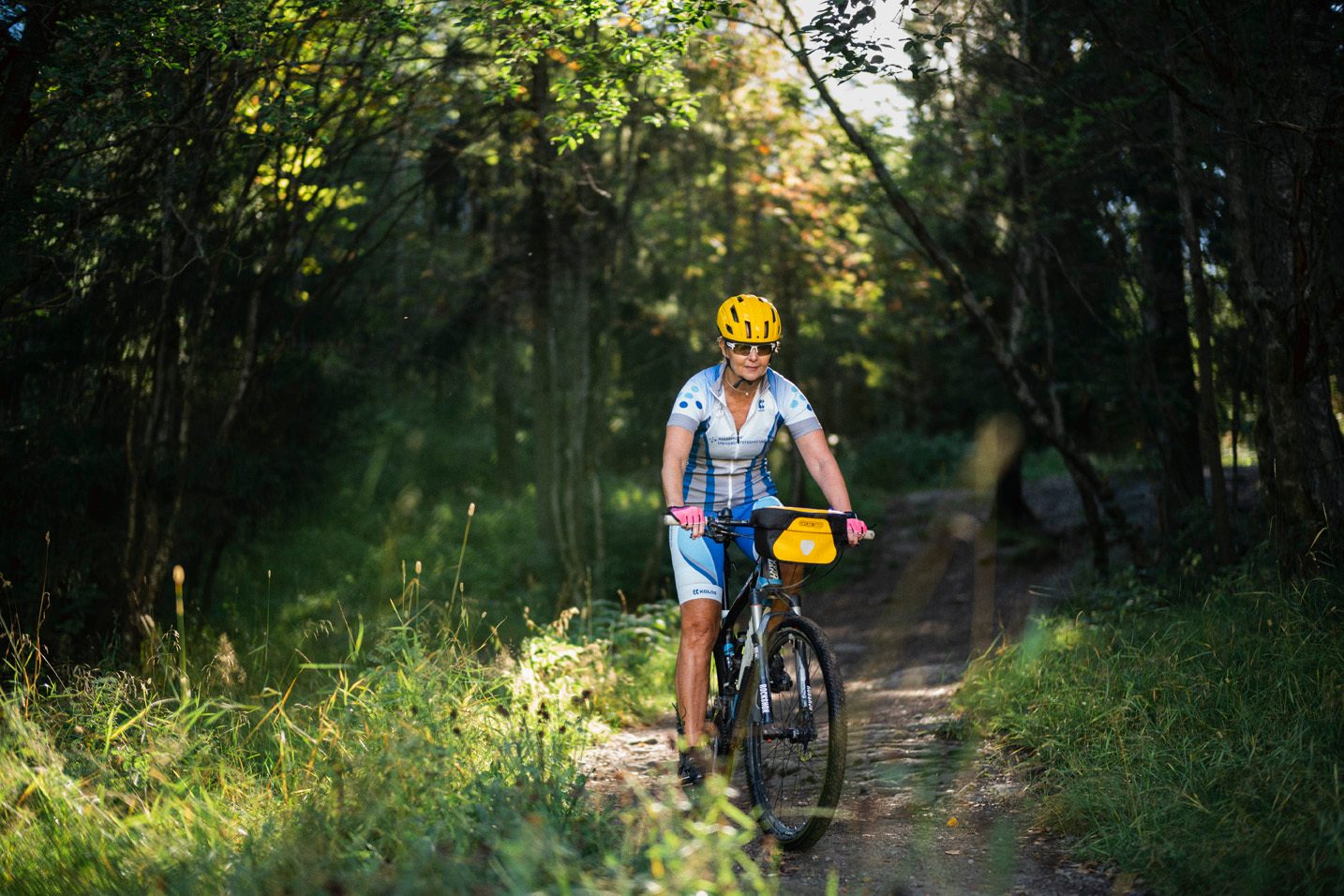 En kvinne i seksti&#xE5;rene sykler i skogen med gul hjelm og raske briller.