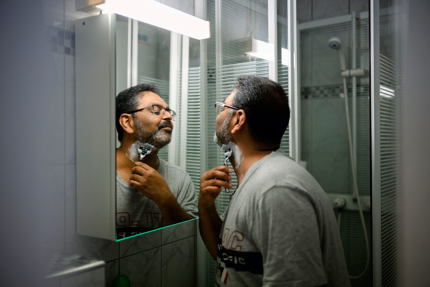 En mann med gr&#xE5; t-skjorte barbererer seg. Vi ser badet og speilet.