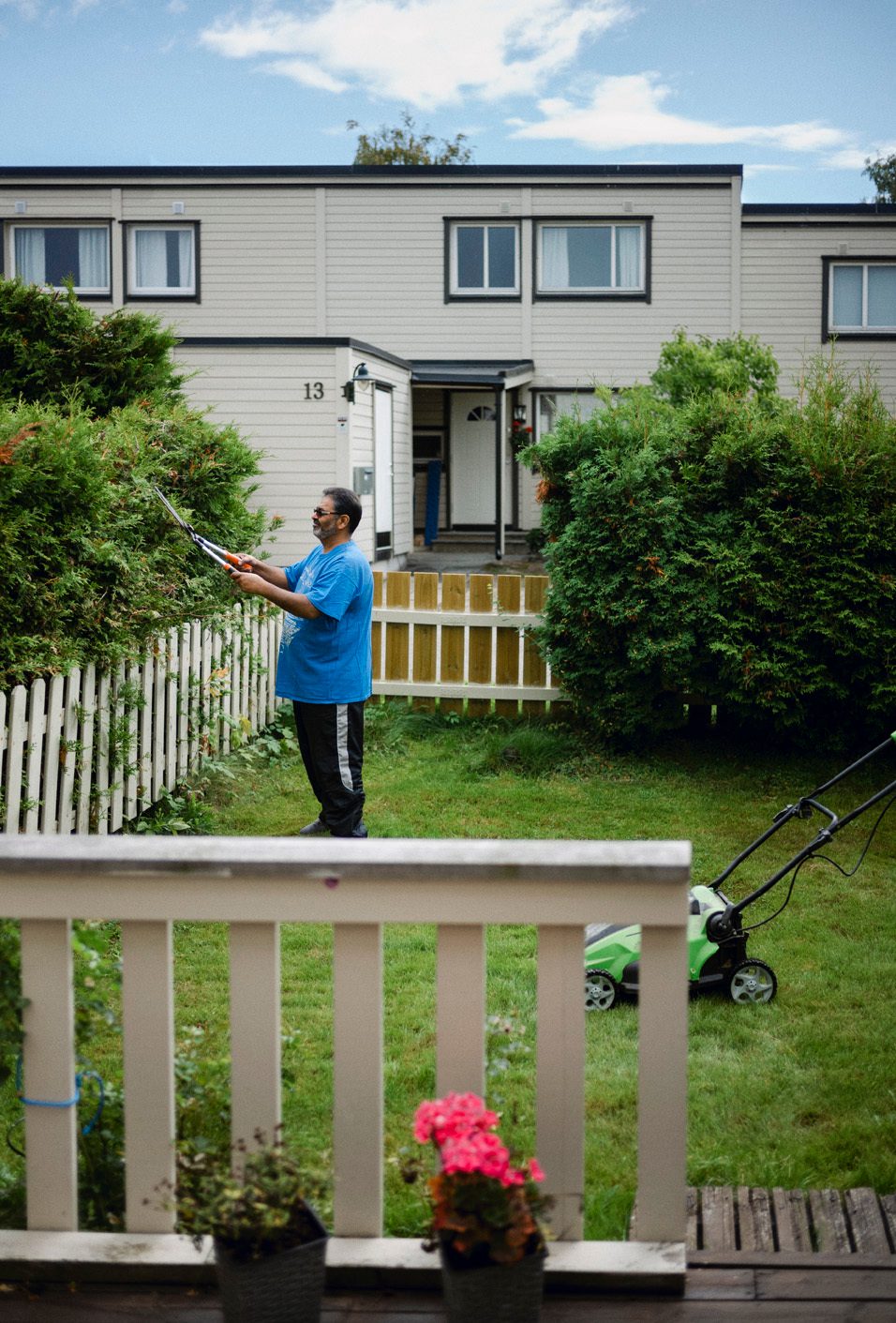 En mann med lysebl&#xE5; t-skjorte klipper hekken i rekkehushage. Det st&#xE5;r en gressklipper p&#xE5; plenen.
