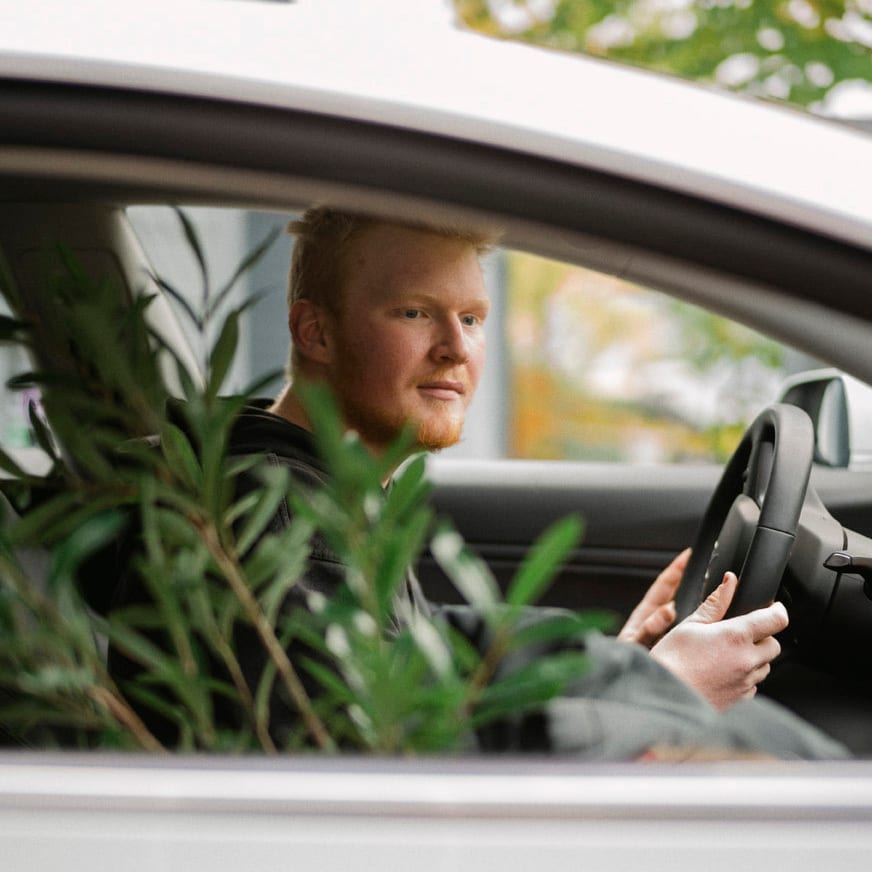 Bilde av en ung mann som kj&#xF8;rer bil med en flyttekasse og gr&#xF8;nn plante i passasjersetet. Bildet er tatt gjennom vinduet.