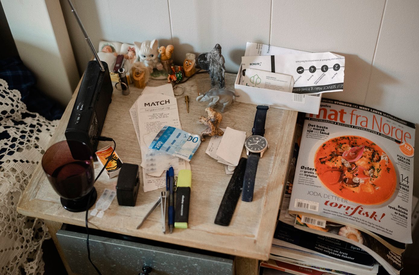 Nattbord til en eldre mann, med radio, fyrstikkeske, kvitteringer, klokke, penner, blader og annet sm&#xE5;rusk.