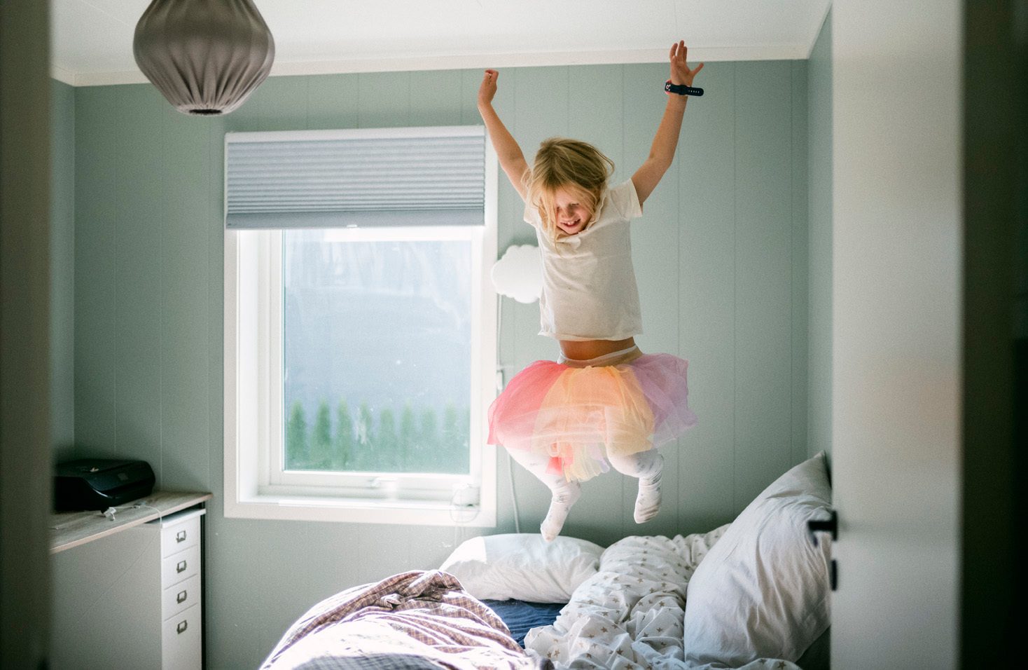 Jente med fargerikt skj&#xF8;rt og hvit t-skjorte hopper i sengen til foreldrene, lysebl&#xE5;tt soverom.