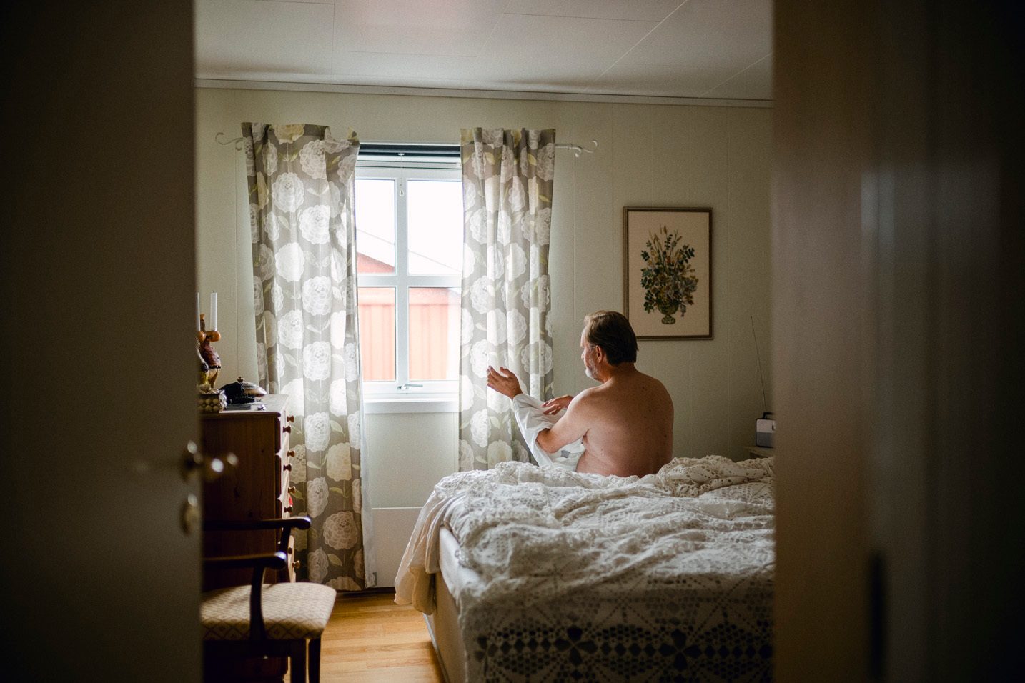 En mann sitter p&#xE5; seng med hvitt heklet sengeteppe og tar p&#xE5; seg skjorte. Bildet er tatt bakfra, med vindu og maleri p&#xE5; veggen