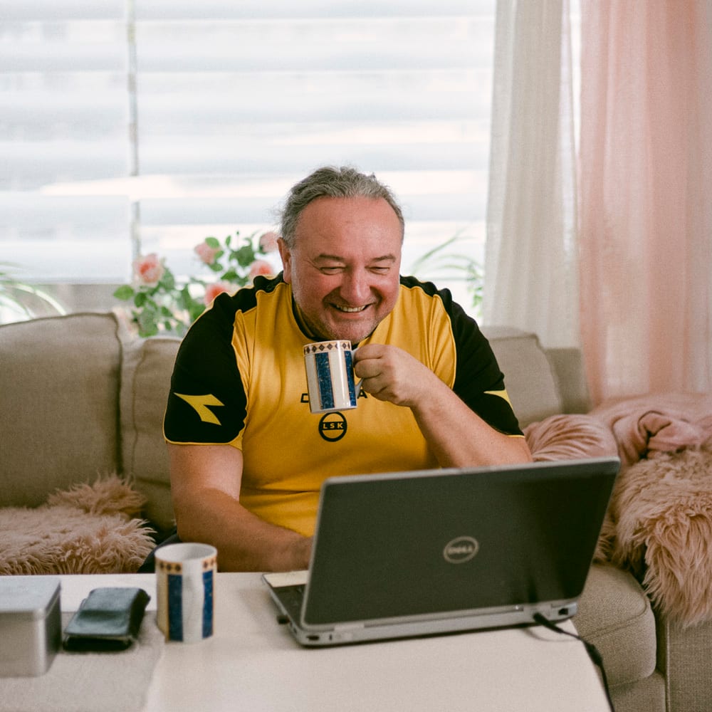En mann i gul fotballtr&#xF8;ye sitter med kaffekopp i beige sofa foran en datamaskin og smiler.