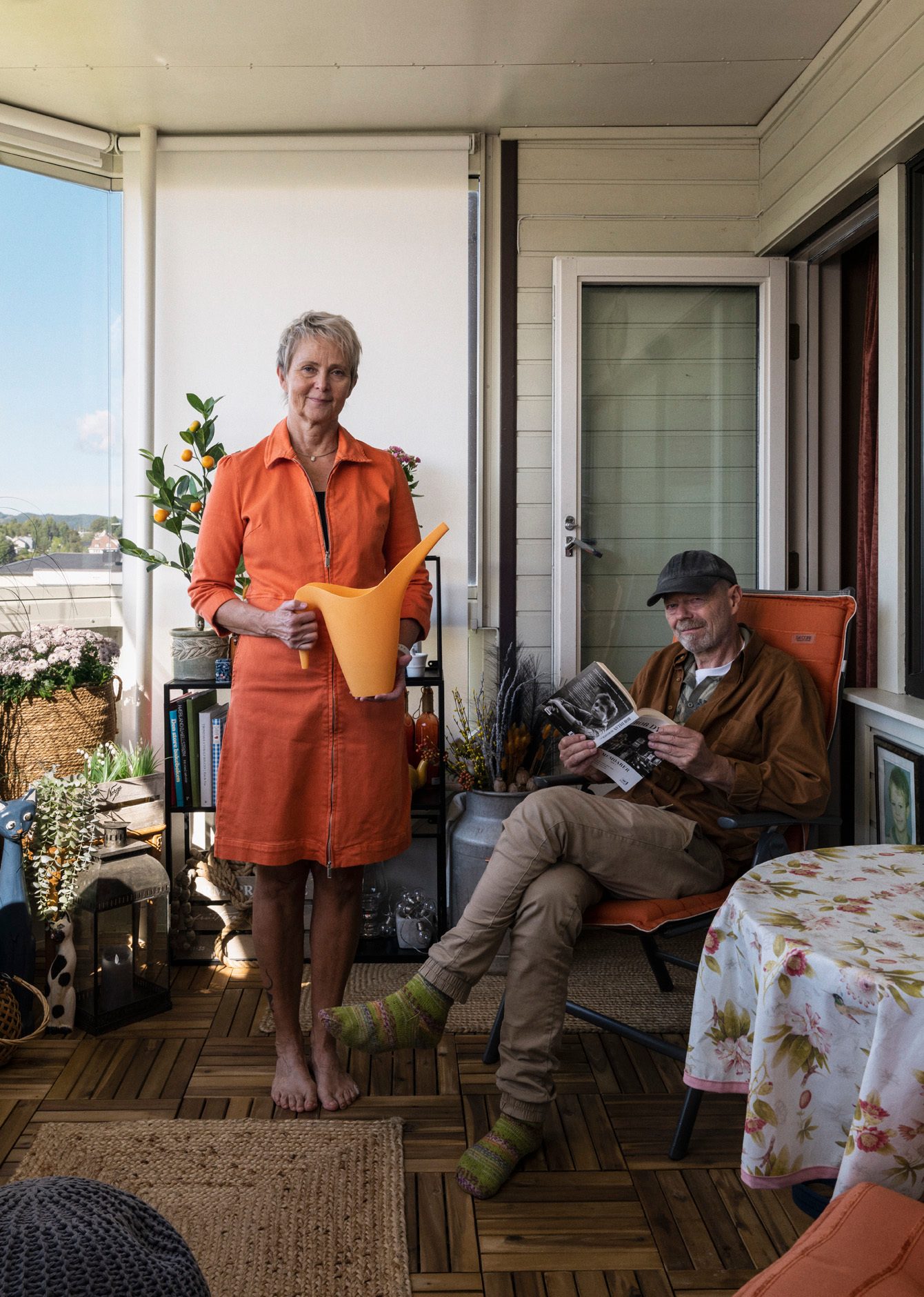 En kvinne st&#xE5;r i oransje kjole med en oransje vannkanne p&#xE5; en innglasset veranda. Ved siden av henne sitter en mann med caps og ullsokker og leser en bok om Bob Dylan.