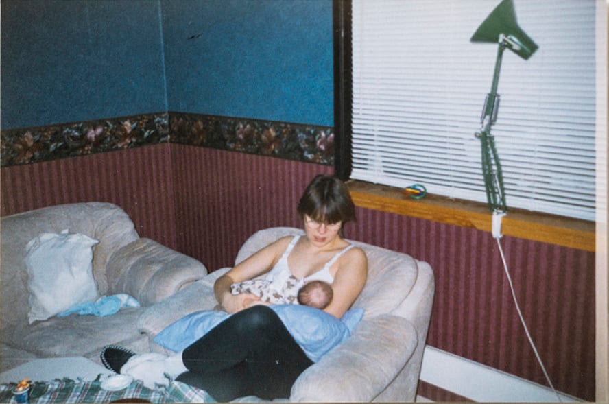 Nybakt mor ammer baby i sofa i en stue med 90-talls interi&#xF8;r. Stuen er opplyst av en skrivebordslampe.
