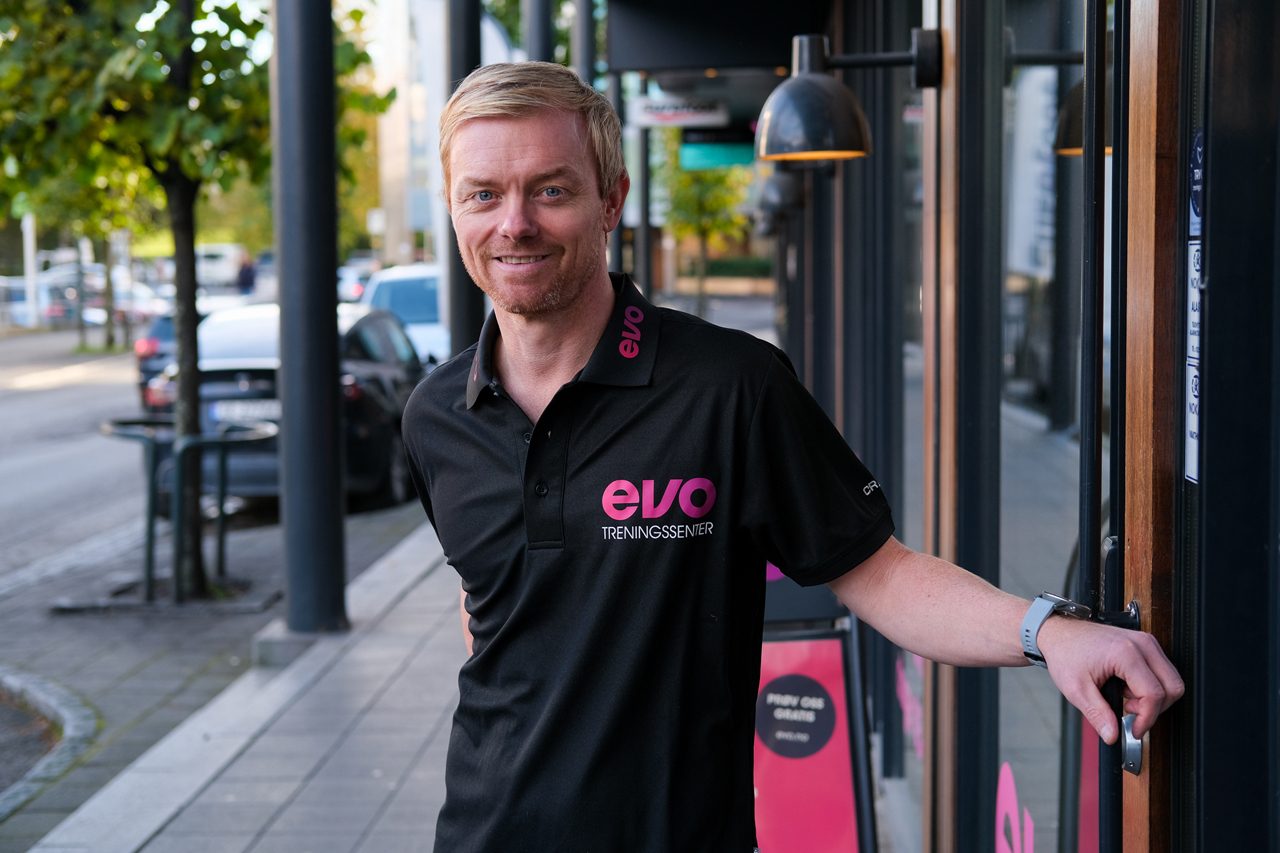 Blond mann med svart polo-skjorte med rosa logo st&#xE5;r utenfor en d&#xF8;r ut mot en trafikkert gate.