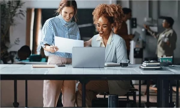 Two smiling women collaborate on a laptop, reviewing documents in a modern office setting.