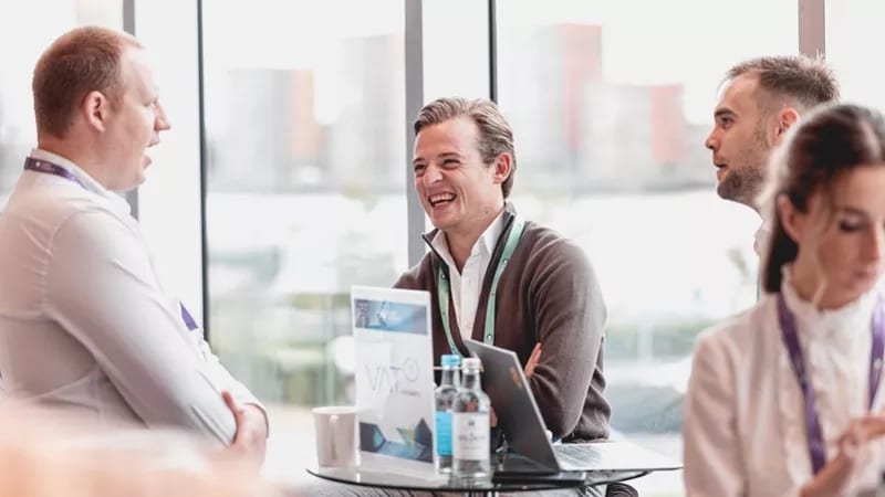 A group of business professionals networking at an event, with one man laughing in the center.