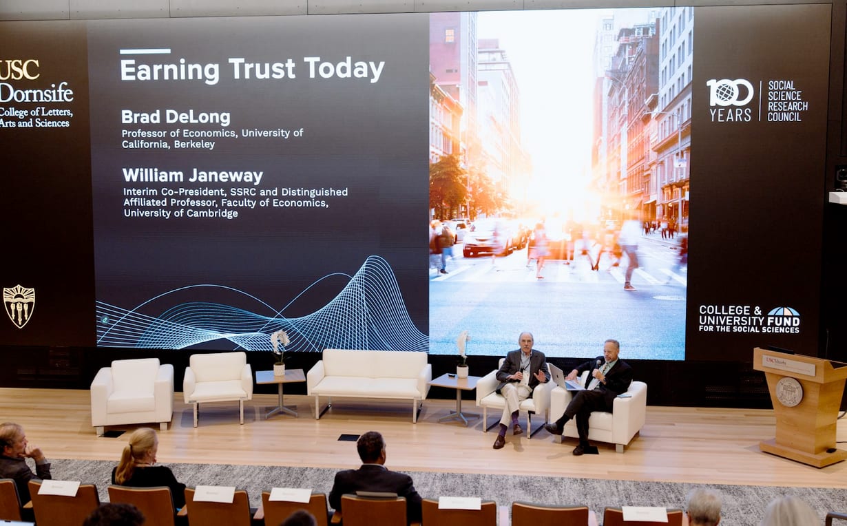 Two speakers on stage in front of a large screen displaying "Earning Trust Today" and a city scene.