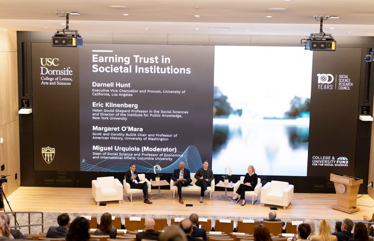 Four panelists discuss 'Earning Trust in Societal Institutions' on stage before an audience, with event details on a large screen.