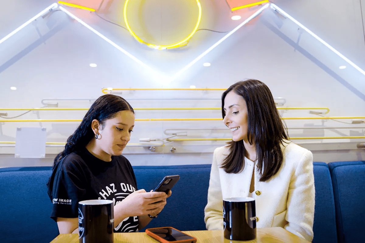 Two women at a table, one looking at her phone, the other smiling, with neon lights in the background.