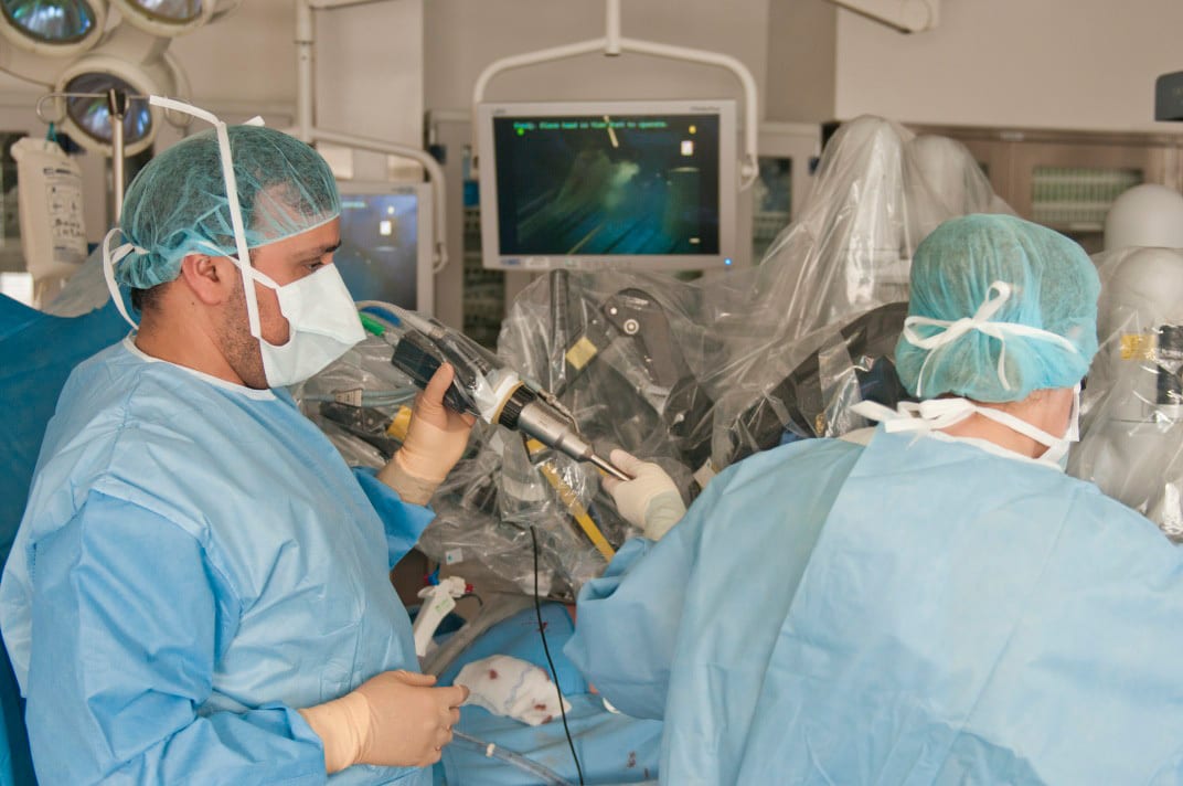 Surgeons perform robotic-assisted surgery in an operating room, viewing the procedure on a monitor.
