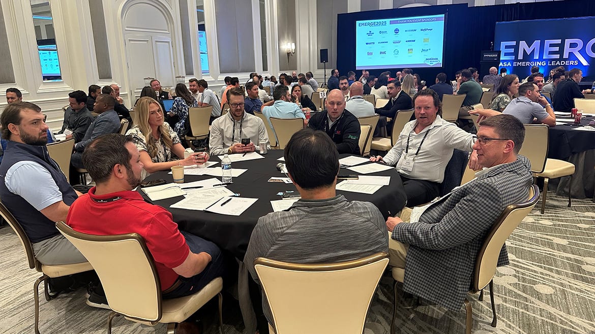 Conference attendees seated at round tables, with a large screen displaying "EMERGE2025" in the background.