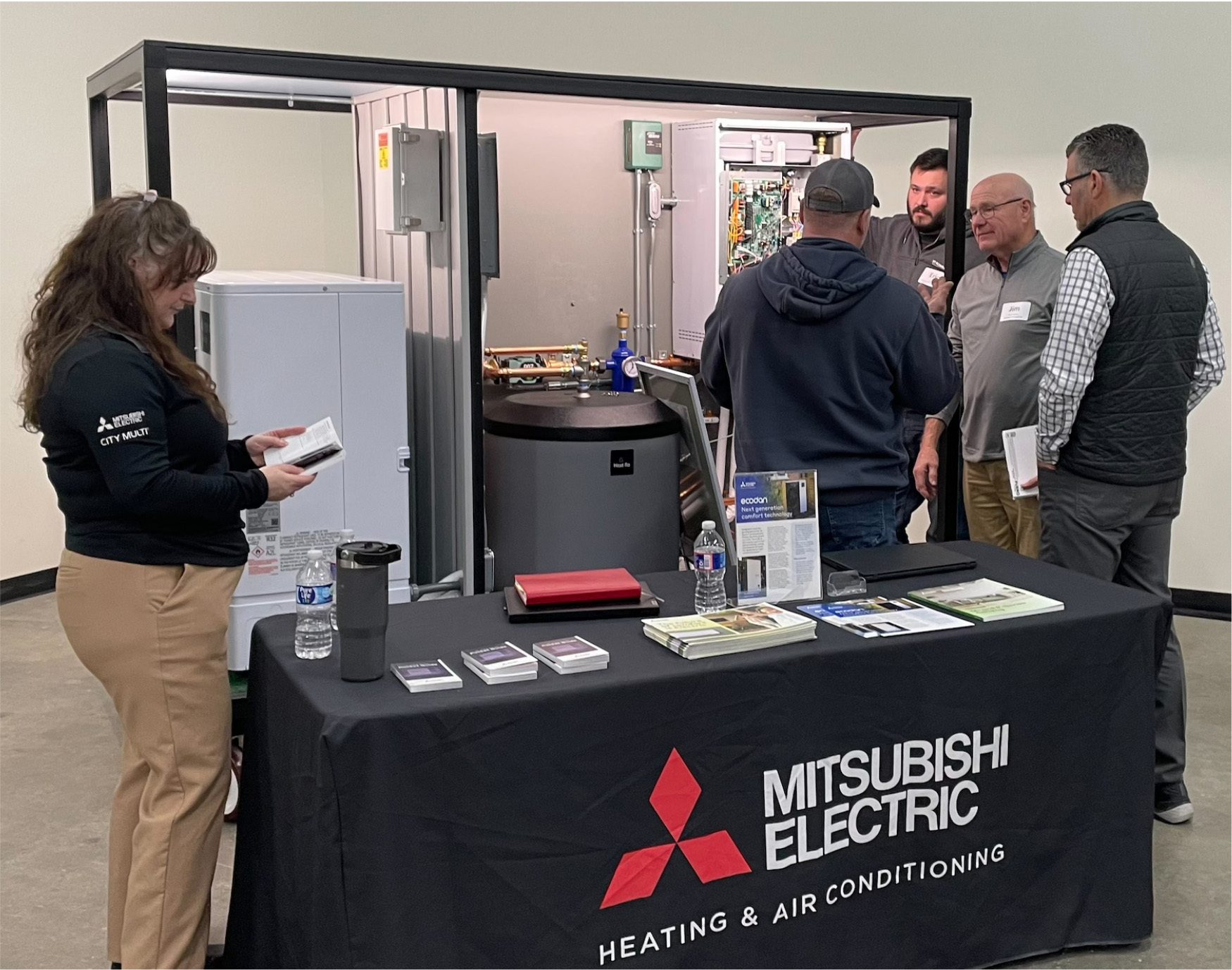 People gather at a Mitsubishi Electric HVAC trade show booth with equipment displays and literature.