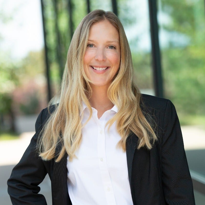 A smiling woman with long blonde hair in a white shirt and black blazer, against a blurred green background.