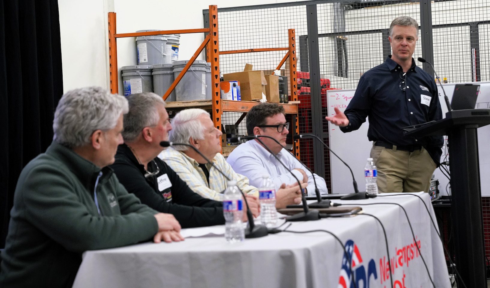 A man speaks at a podium to a panel of four men seated at a table in an industrial-style room.