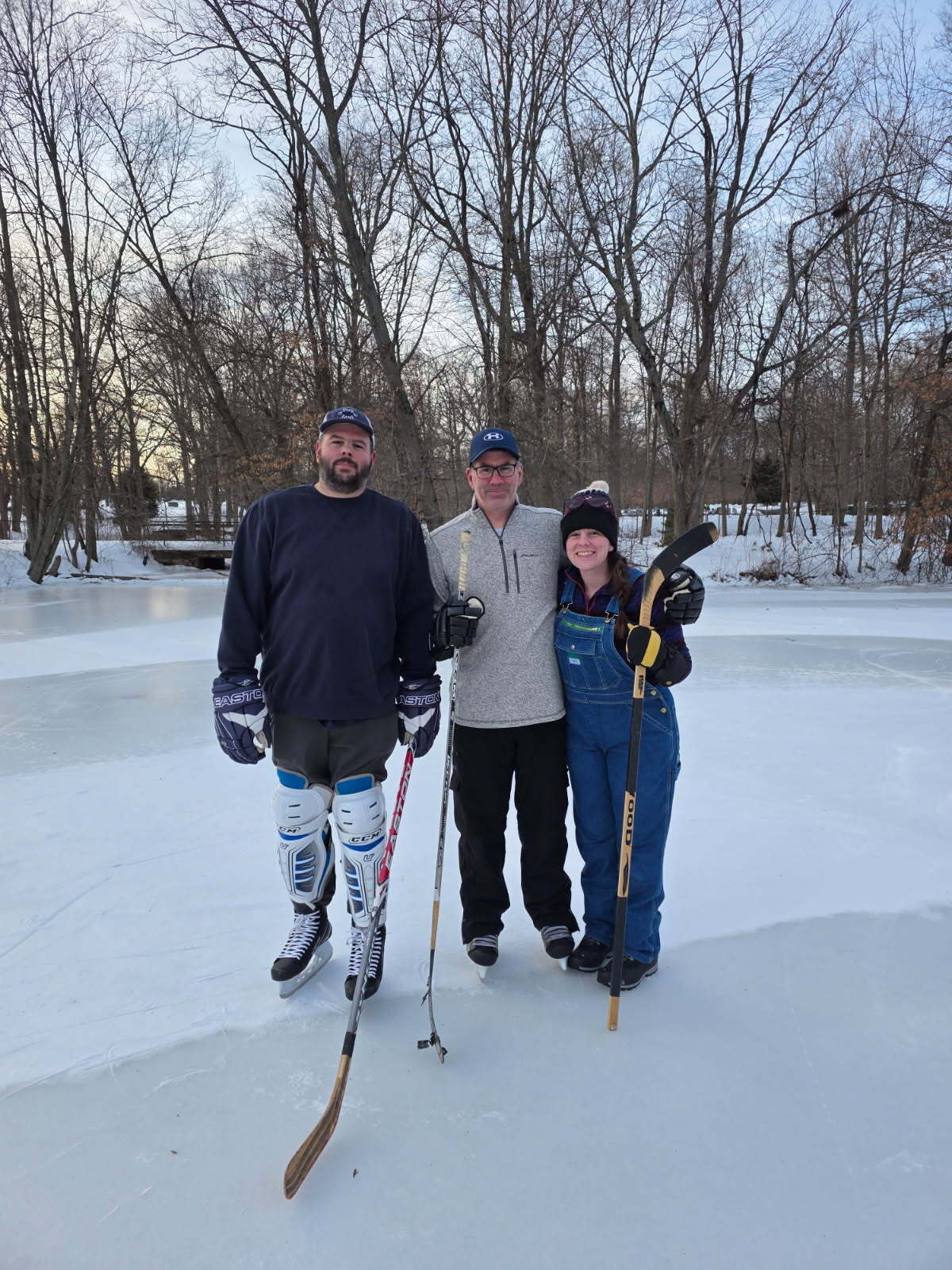 Three people, two men and one woman, with hockey sticks on a frozen pond surrounded by trees.